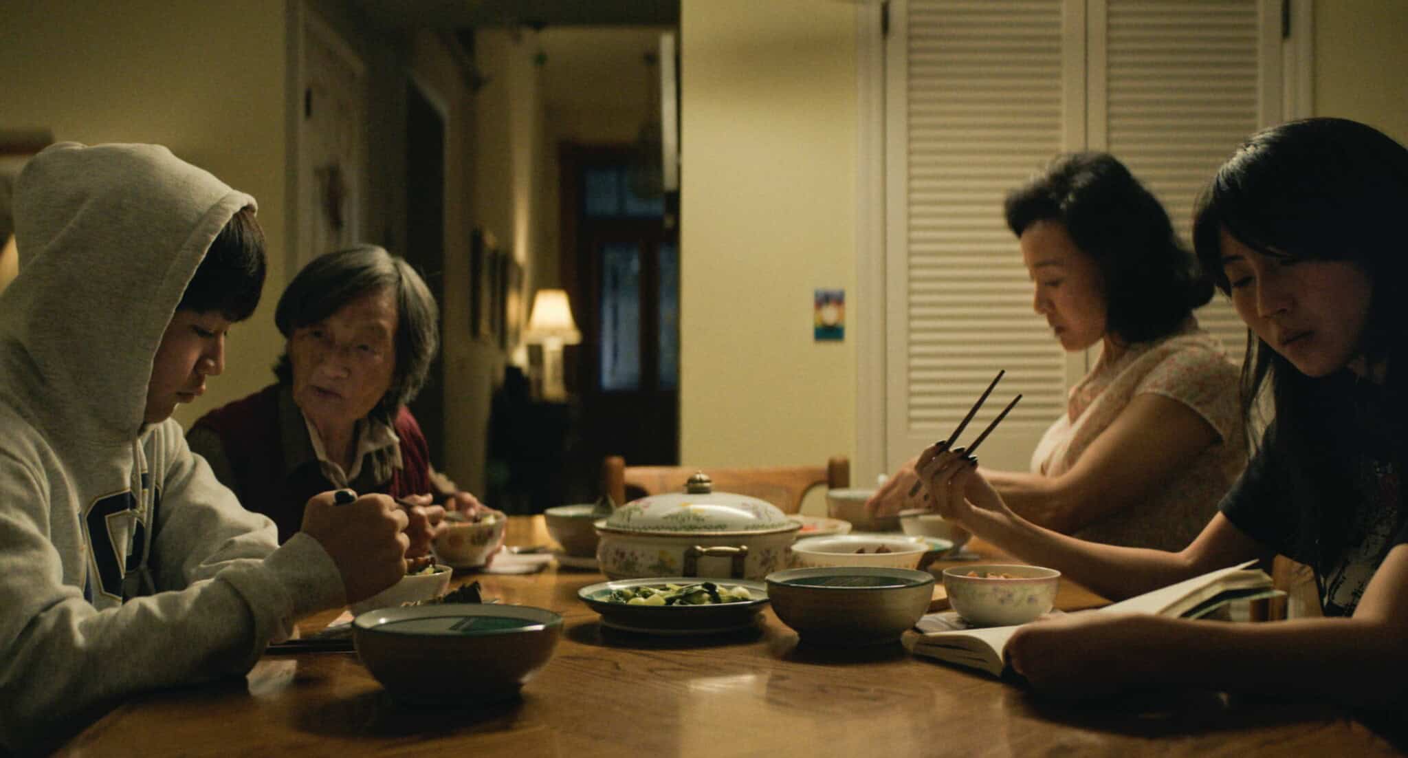 An Asian family, with a mother, grandmother, daughter and son, sit at a dinner table eating.