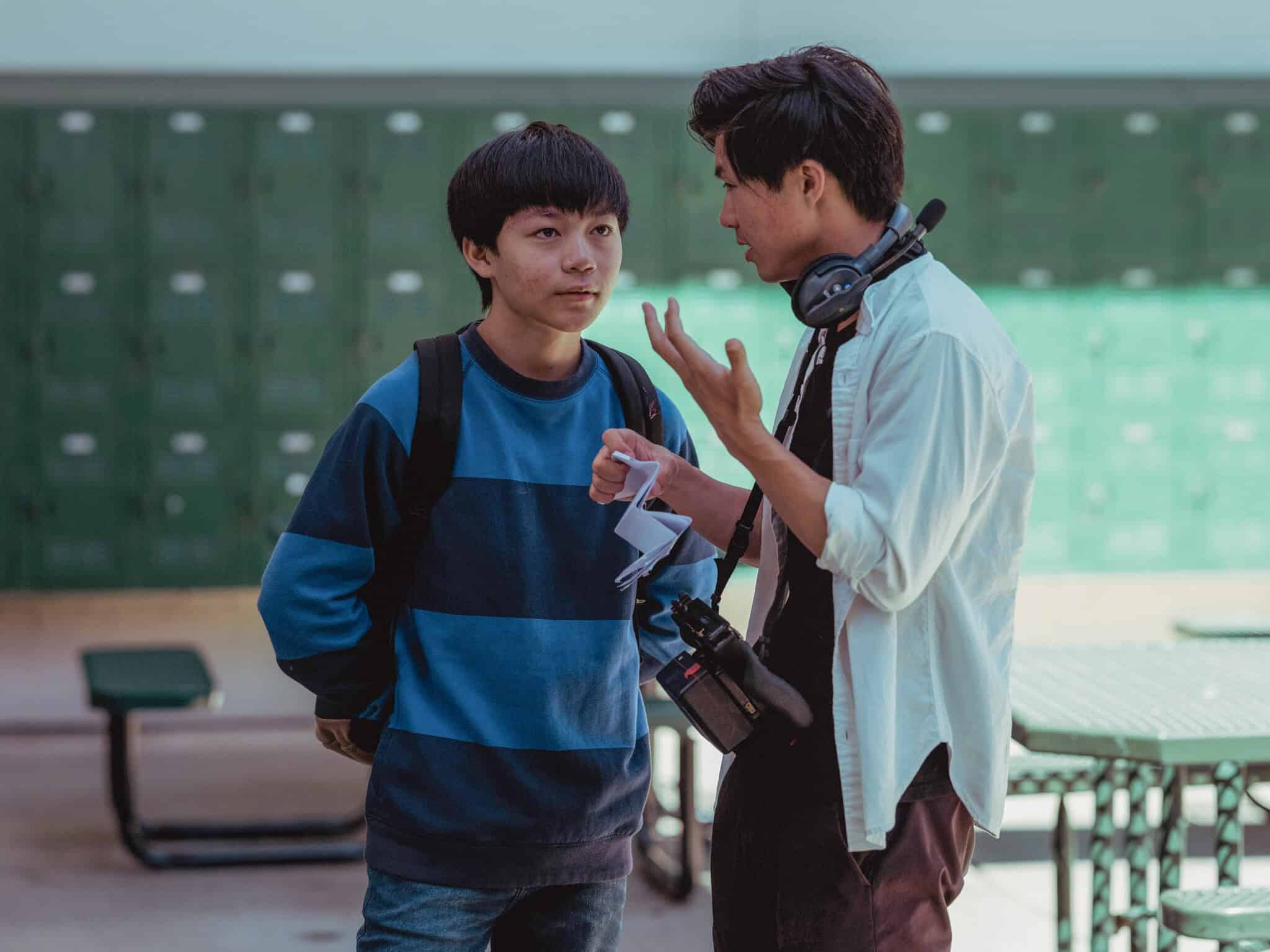 Actor Izaac Wang and director Sean Wang stand talking to each other with lockers and tables in the background on the set of "Didi."