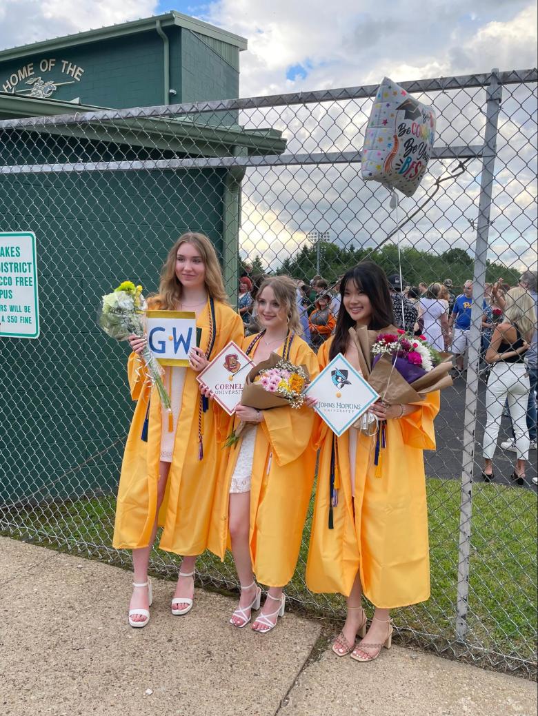 Three people stand against a metal fence wearing yellow graduation gowns.