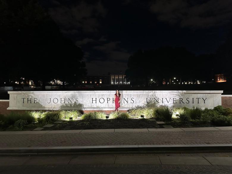 A women in a red dress stands in front of an illuminated wall inscribed with "The Johns Hopkins University."