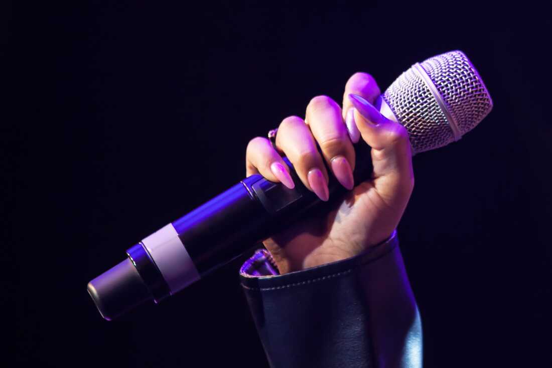 A close-up photograph showing a hand with long, manicured, light pink nails and the wrist of a leather jacket visible, holding a karaoke microphone in a dark room with lights that have a purple cast.