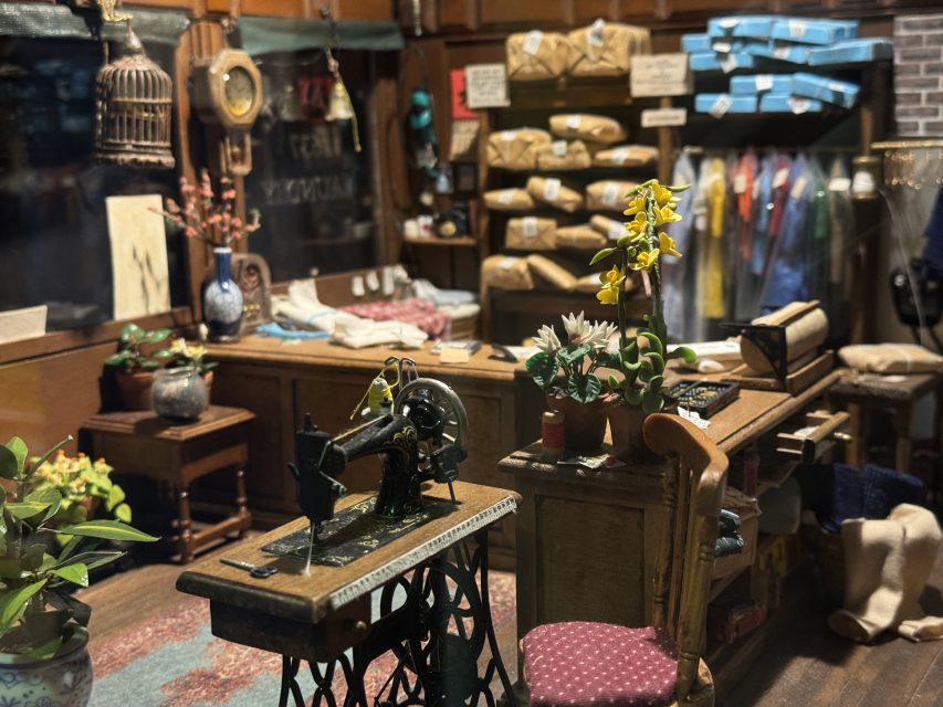A vintage sewing shop interior with a sewing machine, fabric bundles, potted plants, and packages on shelves, with garments hanging in the background.