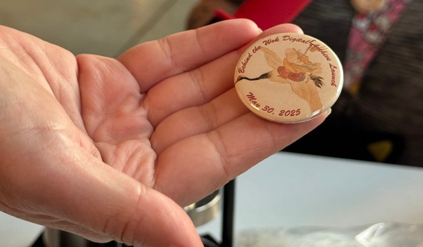A hand holding a commemorative button for the Behind the Wok Digital Archive Launch on May 30, 2025, featuring an illustration and the event name.