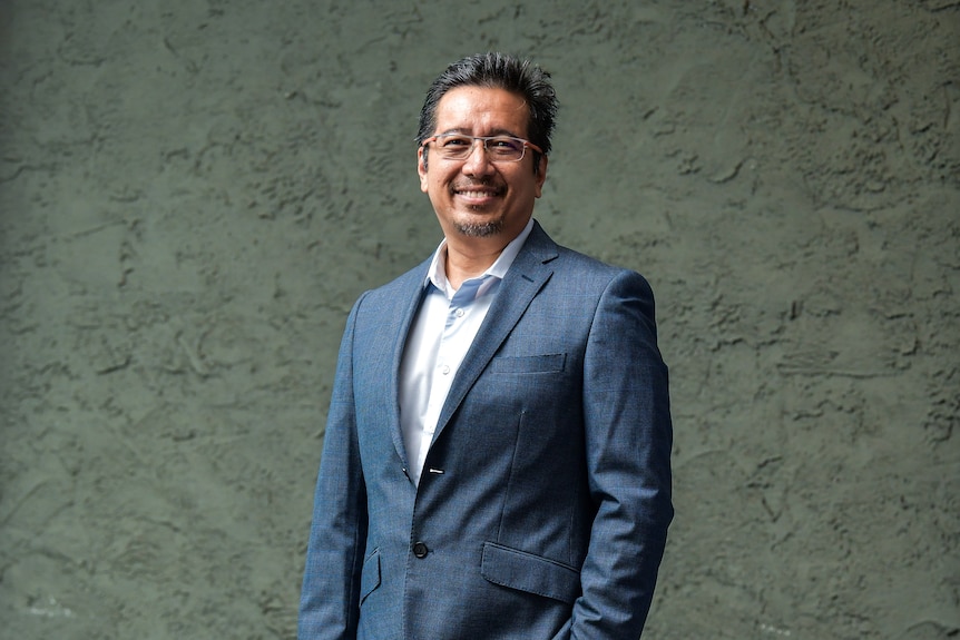 An asian man in a navy blue suit and glasses in front of a dark green background.