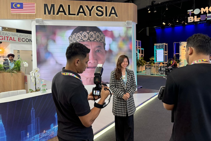 Image shows a girl in a plaid blazer standing in front of two men holding cameras. She's in front of a sign that says 'Malaysia'
