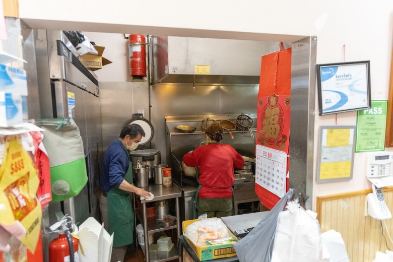 A man and a woman work in the kitchen of a restaurant behind a cluttered counter and a bright red Chinese calendar