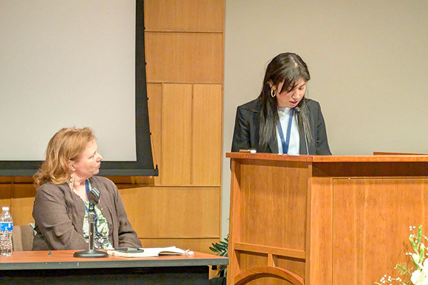 A speaker in a dark blazer addresses an audience from a wooden podium. A woman in a brown cardigan sits nearby, listening.