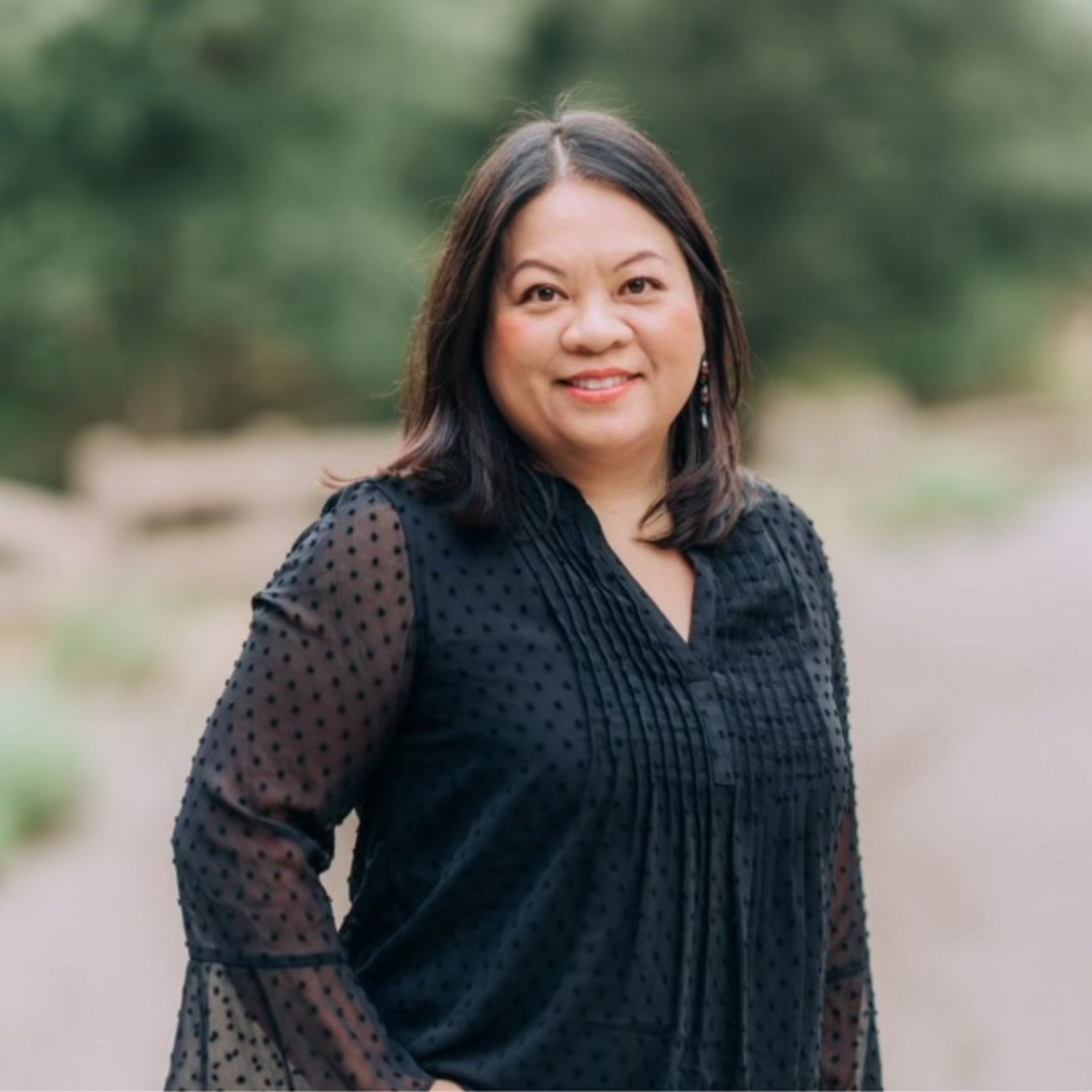 A smiling woman with shoulder-length dark hair wears a sheer black polka-dot blouse, standing outdoors with blurred greenery in the background.