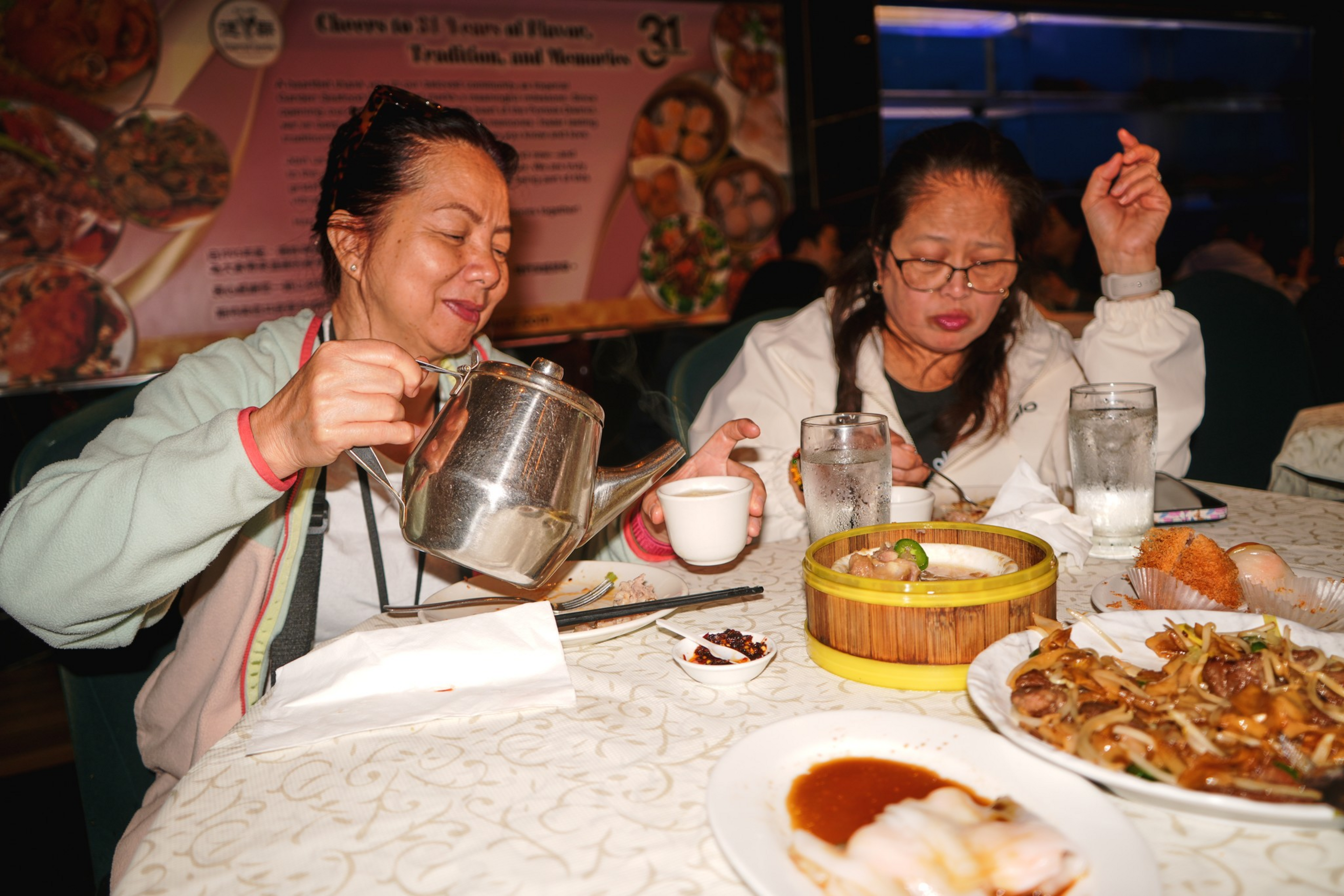 Two women sit at a round table with various Asian dishes; one is pouring tea from a metal teapot into a small cup while the other looks down.