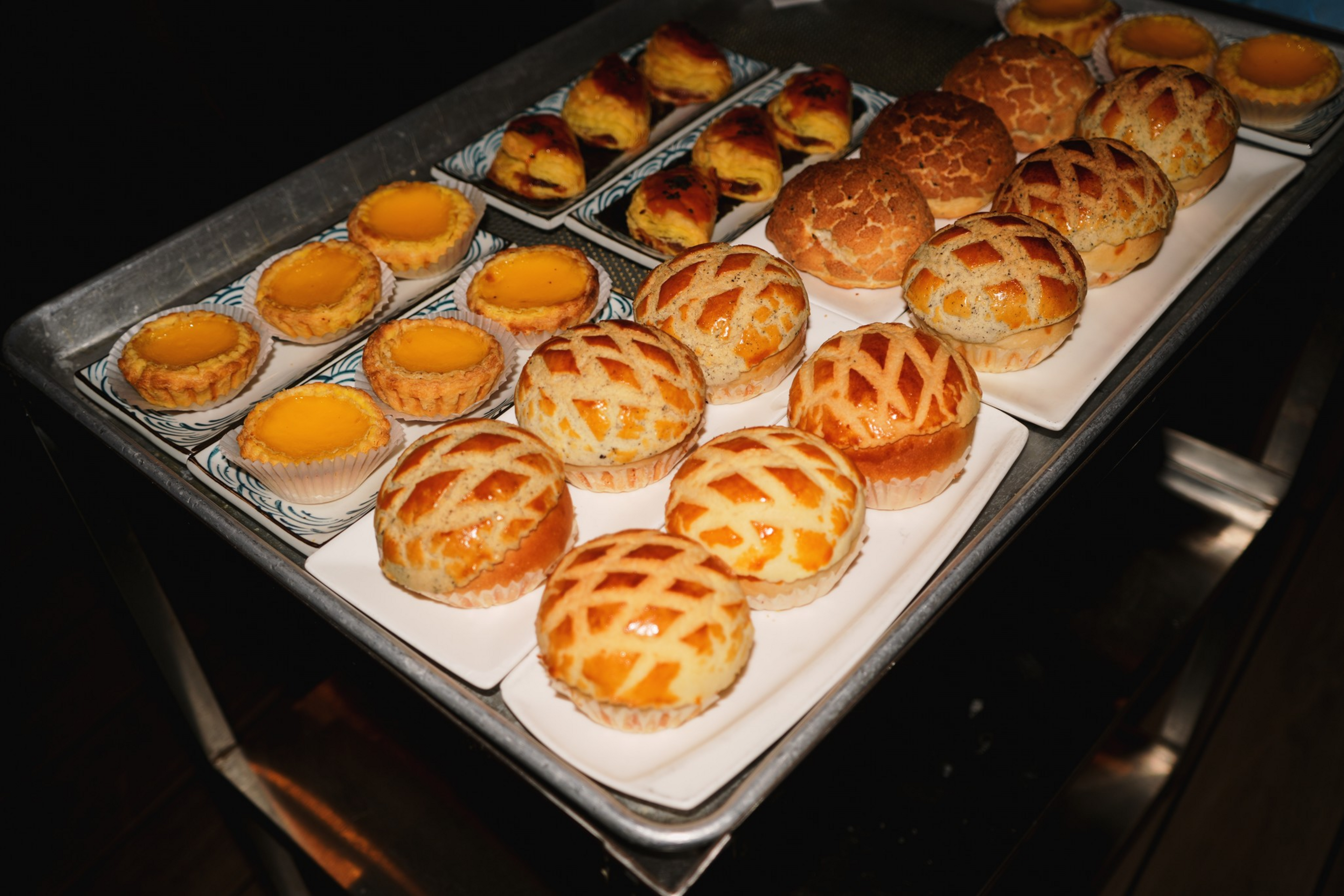 Several trays hold an assortment of baked goods, including egg tarts, pineapple buns with a crisscross pattern, and pastries with a golden, flaky crust.