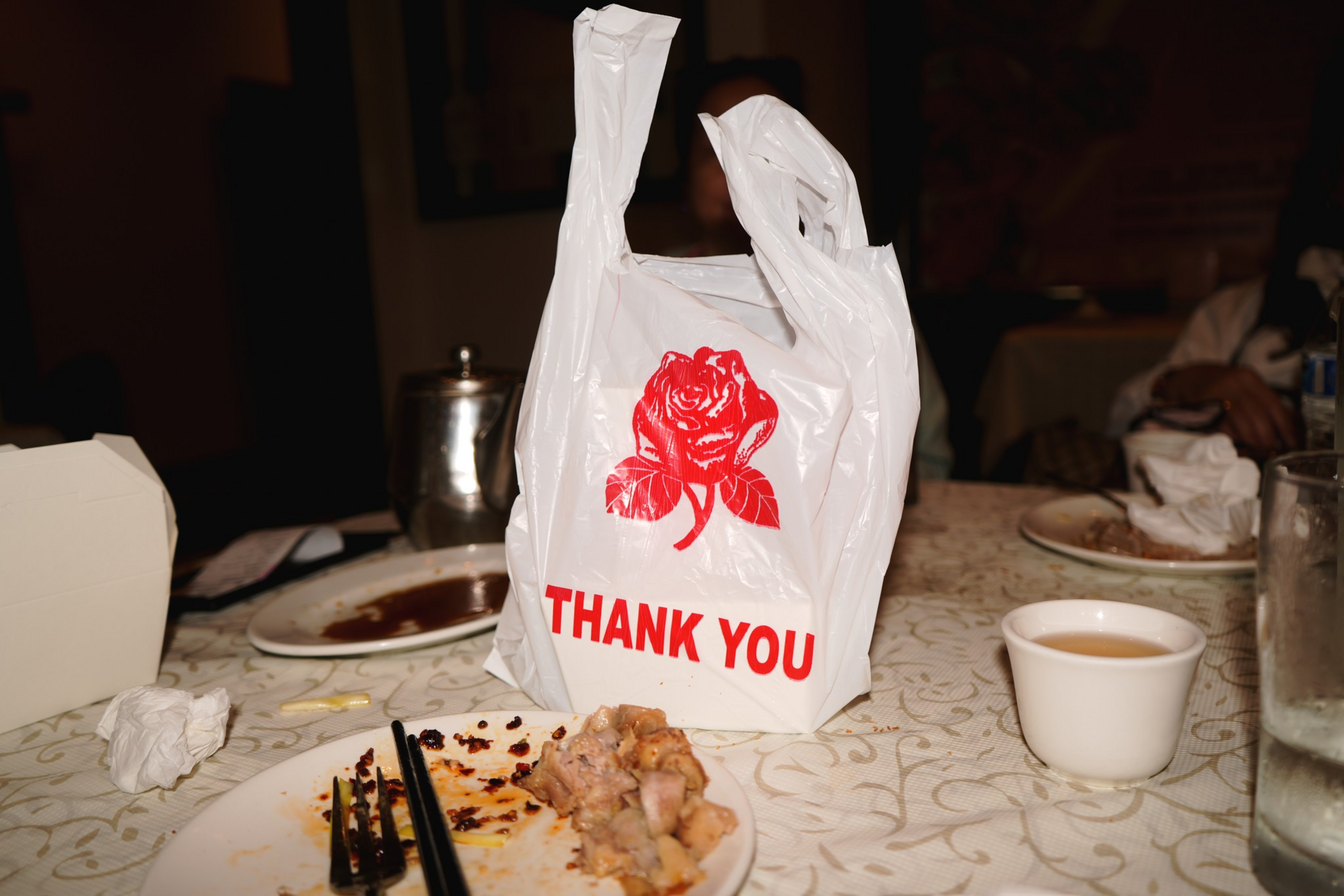 A white plastic takeout bag with a red rose and “THANK YOU” in red sits on a table with empty plates, chopsticks, a cup of tea, and a glass of water.