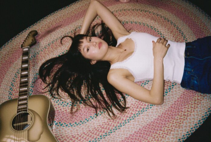 A young Asian woman with long dark hair, in a white tank top, lays on a pink and teal rug, with one arm behind her head and a guitar next to her.