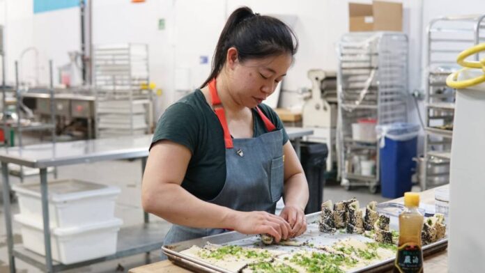 In a bakery kitchen, an Asian woman rolls up pieces of dough covered with chopped scallions.