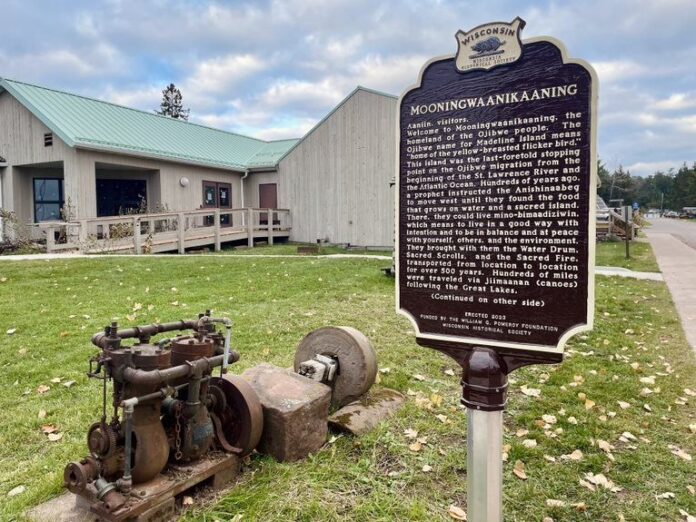 A historic marker about Mooningwanekaaning stands near an old rusted machine outside a beige building with a green roof and a sidewalk on a cloudy day.