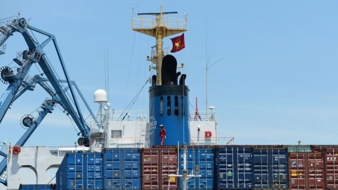 A man walks on top of containers on board a big ship. A large crane is in the background